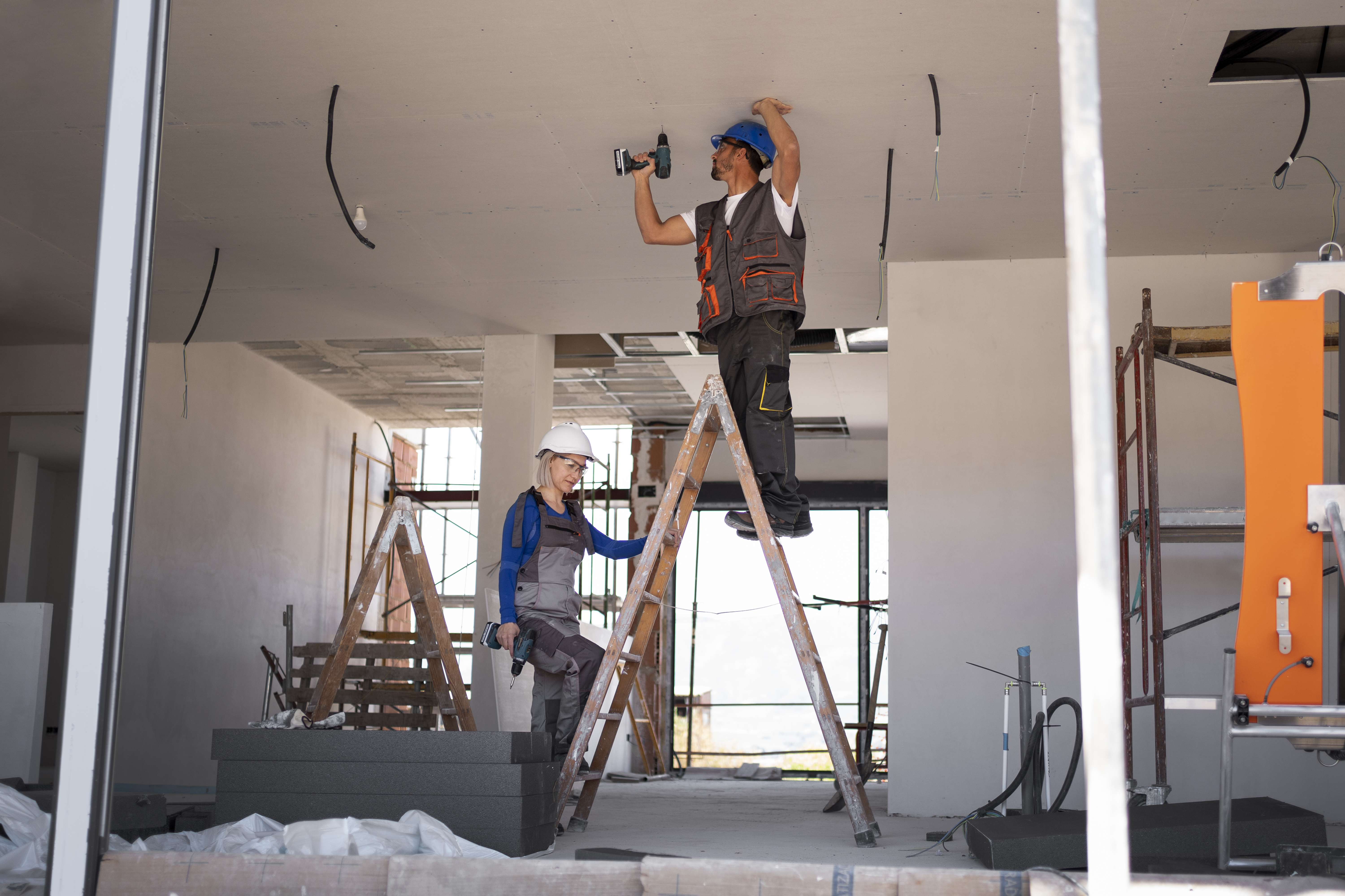 Two construction workers, a man and a woman, standing on separate ladders while working on the ceiling of an unfinished building interior.
