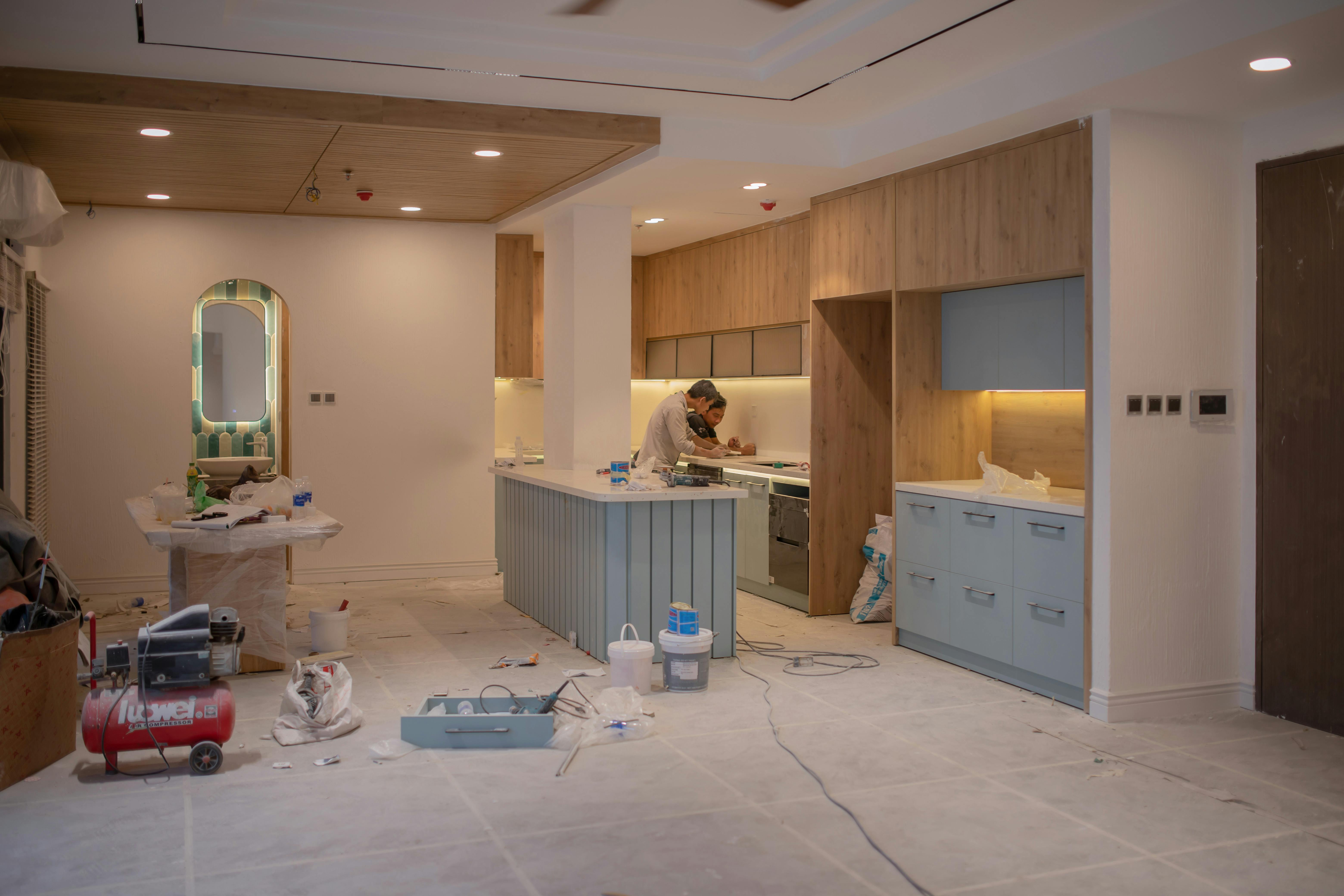 Two workers renovating a modern kitchen space with tools and materials scattered throughout the room.