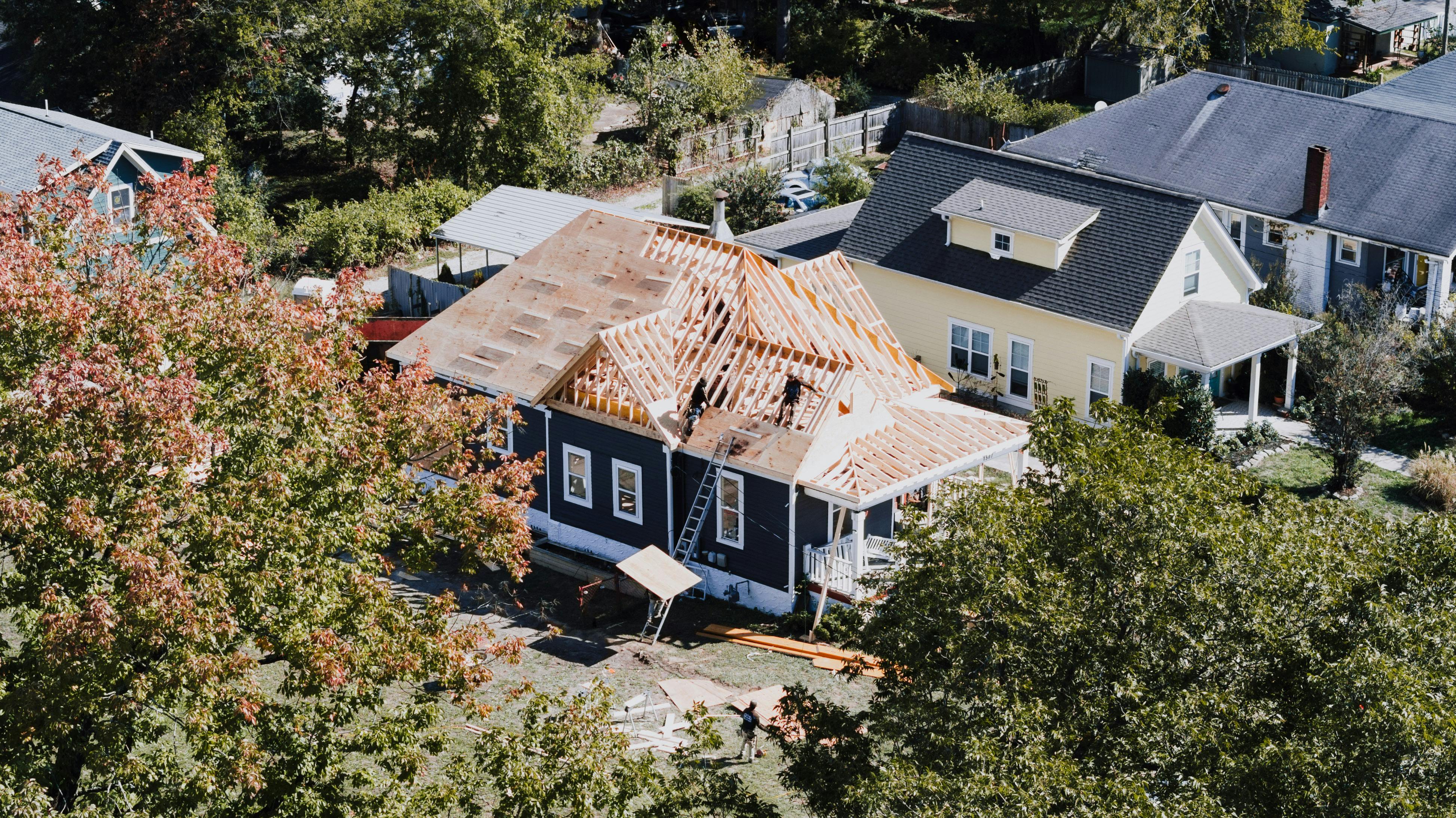 Aerial view of a home under construction with exposed roof framing and workers actively building the new structure.