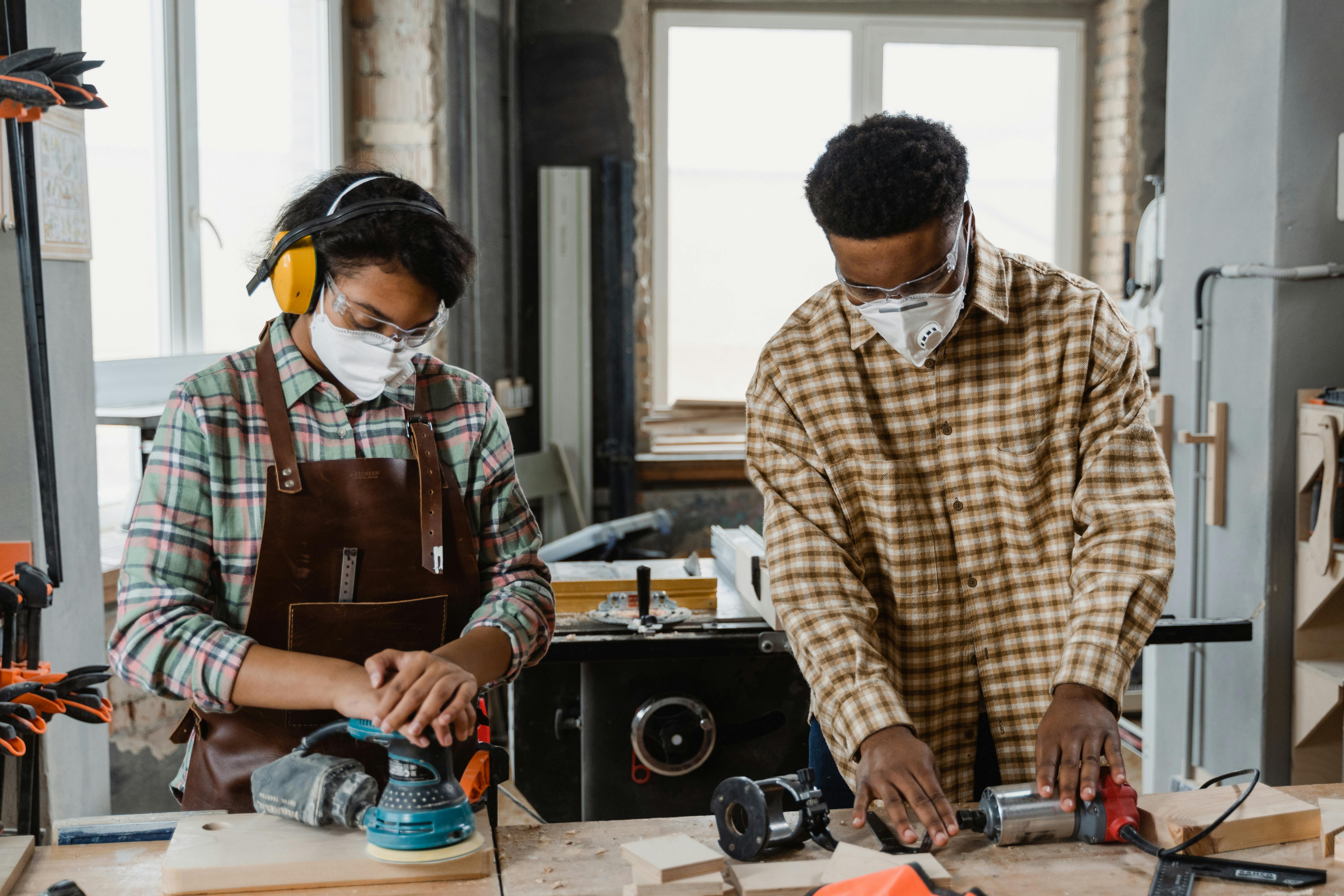 Two individuals wearing protective gear using power tools to sand and drill wood during a home improvement project indoors.